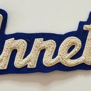 May include: A blue felt patch featuring the name Annette in white, textured, chenille-style script lettering. The patch has a die-cut shape that follows the contour of the cursive text, creating a bold contrast between the white letters and blue base.
