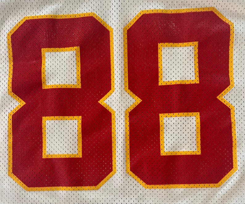 Op de afbeelding: Close-up van een wit mesh-shirt met het nummer 88 in rood met een gele omlijning. De cijfers zijn groot en gecentreerd. Het shirt heeft een geperforeerde textuur.