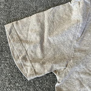 Pu&ograve; includere: Primo piano di una maglietta grigio melange a maniche corte. L'immagine si concentra sulla manica e sulla spalla, mostrando la trama del tessuto e i dettagli delle cuciture. La maglietta sembra essere realizzata in un materiale morbido e confortevole.