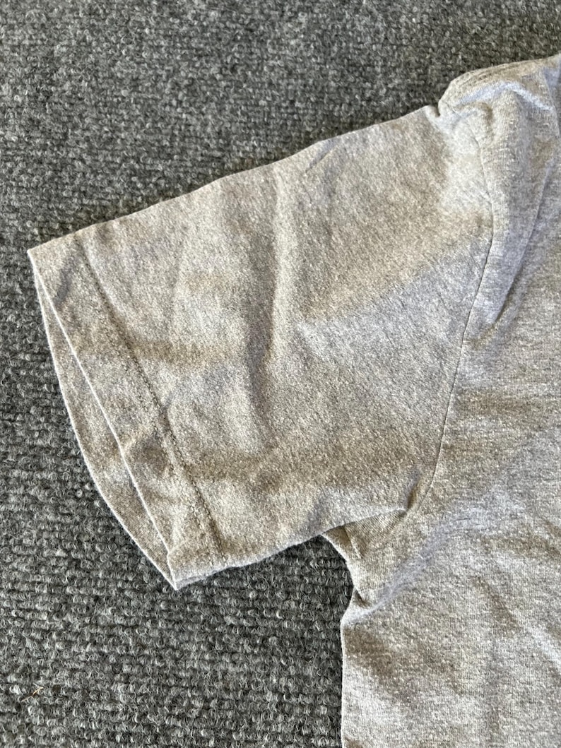 Pu&ograve; includere: Primo piano di una maglietta grigio melange a maniche corte. L'immagine si concentra sulla manica e sulla spalla, mostrando la trama del tessuto e i dettagli delle cuciture. La maglietta sembra essere realizzata in un materiale morbido e confortevole.