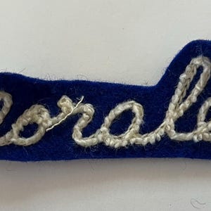 May include: A rectangular blue felt patch with rounded edges featuring the name Morales embroidered in white cursive thread. The embroidery follows the contour of the letters, creating a raised texture against the smooth, dark blue fabric background.
