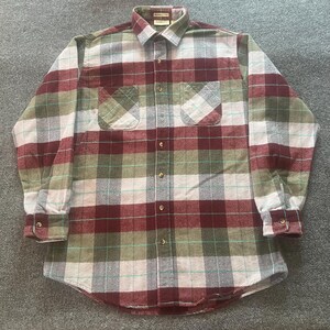 May include: A long-sleeve button-down shirt featuring a plaid pattern in shades of maroon, olive green, and white with thin teal horizontal and vertical lines. The shirt includes a standard collar, two chest pockets, and brown buttons.