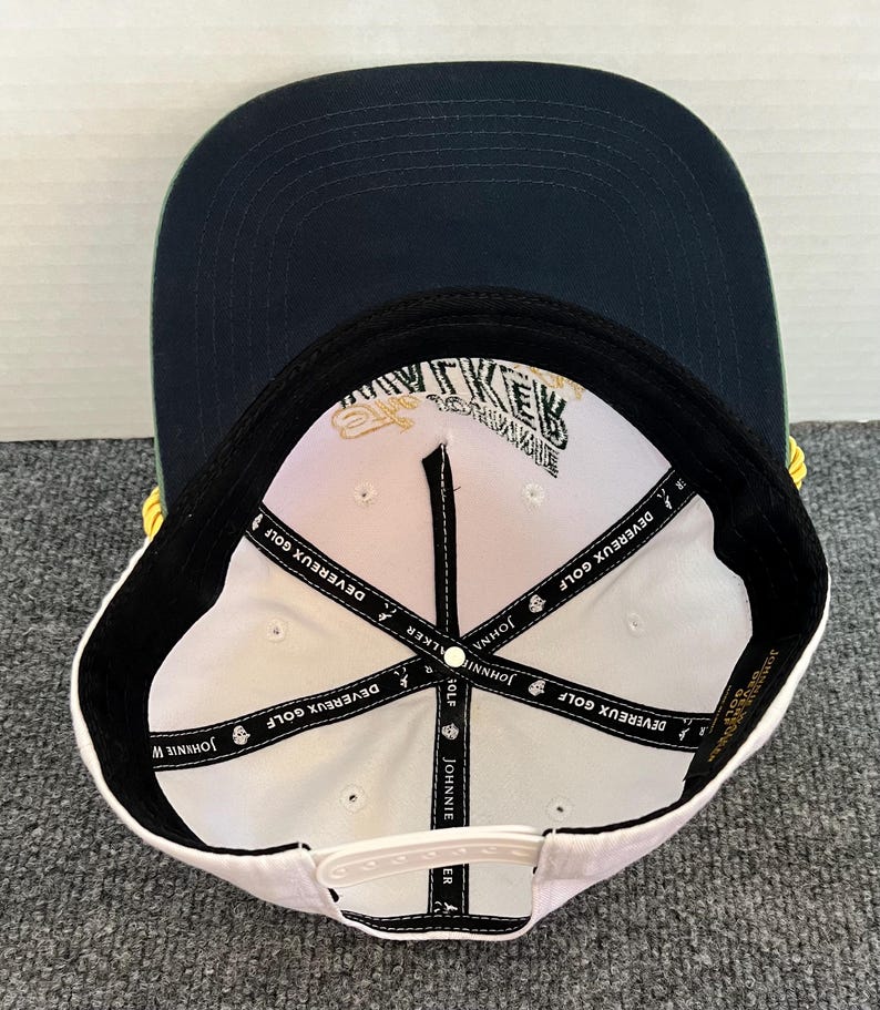May include: A navy blue and white baseball cap with the text "JOHNNIE WALKER" and "DEVEREUX GOLF" printed on the inside. The cap has a flat brim and a white snapback closure. The cap is a golf accessory.