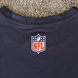 Op de afbeelding: Marineblauw T-shirt met het NFL-logo op de borst. Het logo heeft de vorm van een schild met een voetbal en sterren, en de letters "NFL" in rood. Het T-shirt heeft een ronde hals.