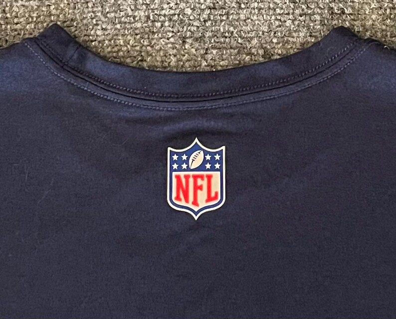 Op de afbeelding: Marineblauw T-shirt met het NFL-logo op de borst. Het logo heeft de vorm van een schild met een voetbal en sterren, en de letters "NFL" in rood. Het T-shirt heeft een ronde hals.