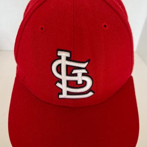 May include: A red baseball cap with a white embroidered logo. The logo is a stylized letter "S" and "L" intertwined. The cap has a curved brim and a rounded crown. The cap is made of a soft, durable material.
