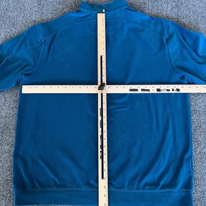 May include: A teal blue long-sleeved shirt with a mock neck and zipper closure. The shirt is laid flat on a gray surface with a wooden ruler positioned vertically and horizontally across the shirt.