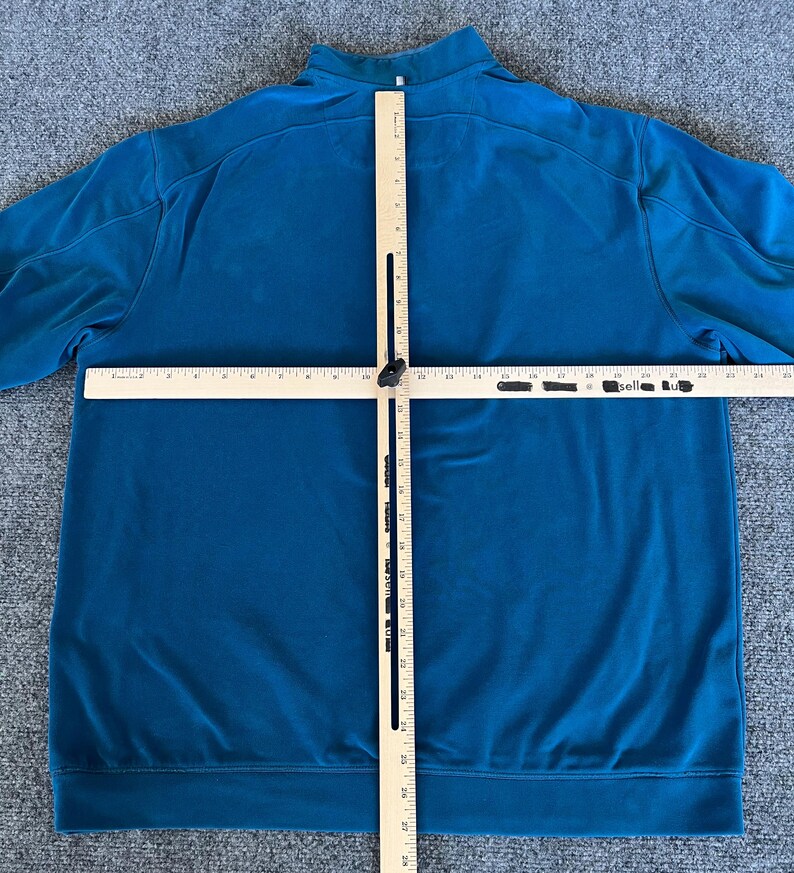May include: A teal blue long-sleeved shirt with a mock neck and zipper closure. The shirt is laid flat on a gray surface with a wooden ruler positioned vertically and horizontally across the shirt.