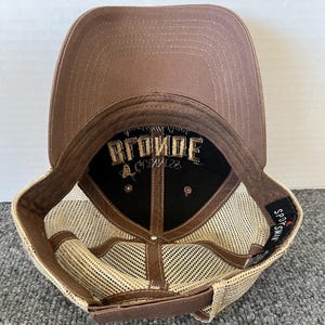 Op de afbeelding: Bruine baseballcap met een mesh achterkant en een gebogen klep. De binnenkant heeft het woord "BLONDE" geborduurd in gouddraad op een zwarte achtergrond. De cap heeft een druksluiting.