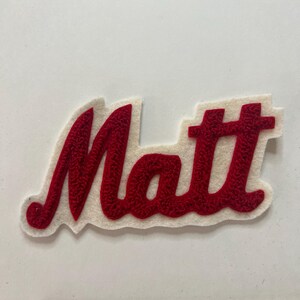 May include: A red embroidered patch featuring the name Matt in a cursive script font, set against a white felt backing. A wooden ruler is positioned at the bottom, showing the patch measures approximately 6 inches in total width.