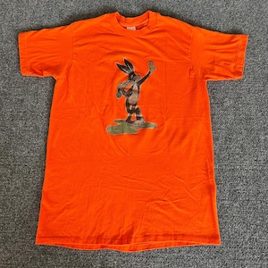 May include: Bright orange t-shirt featuring a cartoon character. The graphic depicts a brown and black rabbit-like figure holding a stick, standing on a green patch. The shirt is a short-sleeved crew neck.