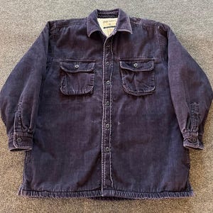 May include: A dark purple long-sleeve corduroy shirt featuring a button-down front, a pointed collar, and two chest pockets with buttoned flaps. The interior is lined with a light-colored fleece material, and the cuffs are secured with buttons.