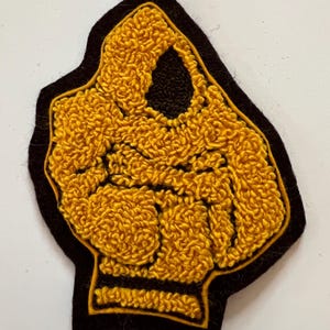 May include: A punch needle patch featuring a raised, textured mustard yellow yarn design of a clenched fist on a solid black felt backing. The design is outlined with a thin mustard yellow yarn border, creating a distinct contrast against the dark background.