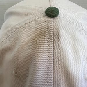 May include: Close-up of a light beige baseball cap with a dark green button on top. The cap has visible stitching and a small air vent. The fabric shows some discoloration.