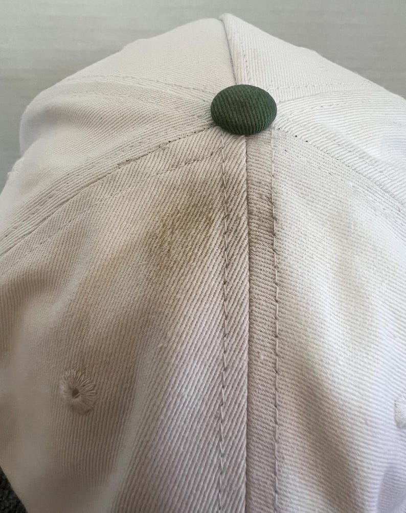 May include: Close-up of a light beige baseball cap with a dark green button on top. The cap has visible stitching and a small air vent. The fabric shows some discoloration.