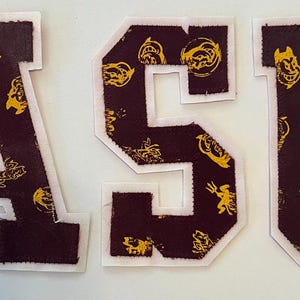 May include: Three fabric letters spelling ASU, each featuring a dark maroon background with a repeating gold pattern of a mascot head and trident. Each letter has a thick white border outline, creating a bold, collegiate-style applique design.