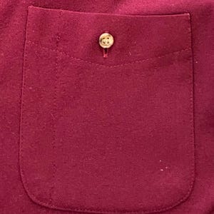 May include: A deep magenta or dark pink fabric pocket featuring a single, centered, light-colored circular button. The pocket has a rounded bottom edge and is stitched onto a matching magenta fabric background with a visible vertical seam line.