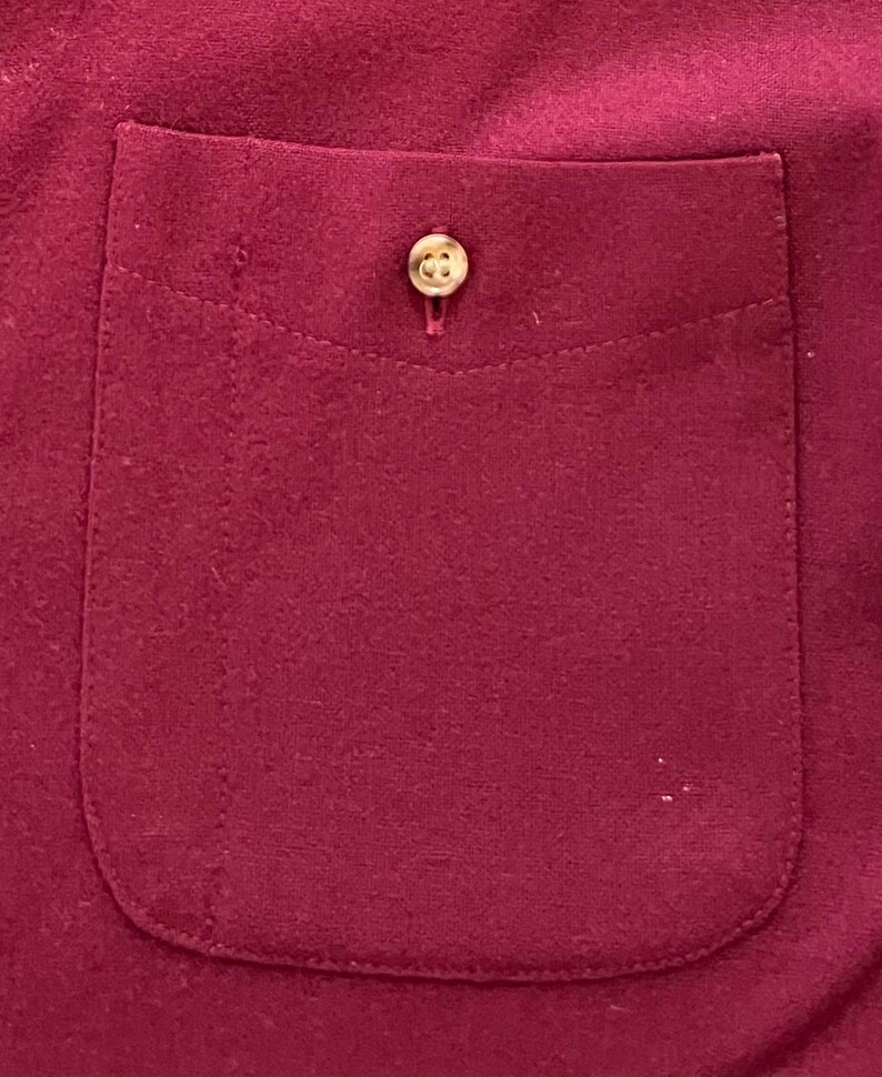 May include: A deep magenta or dark pink fabric pocket featuring a single, centered, light-colored circular button. The pocket has a rounded bottom edge and is stitched onto a matching magenta fabric background with a visible vertical seam line.