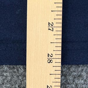 Op de afbeelding: Close-up van marineblauwe stof met een houten liniaal. De liniaal toont metingen in centimeters, met de 68 cm en 71 cm markeringen zichtbaar. De stof lijkt een gebreid of jersey materiaal te zijn.