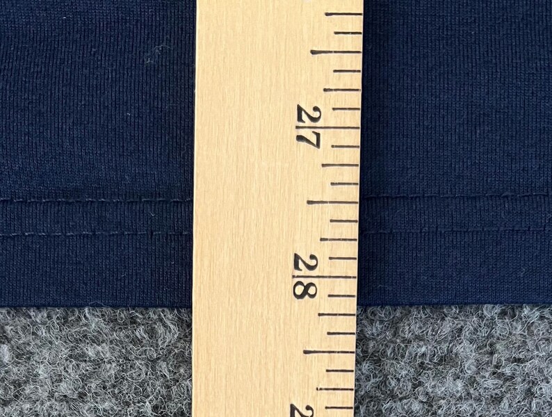 Op de afbeelding: Close-up van marineblauwe stof met een houten liniaal. De liniaal toont metingen in centimeters, met de 68 cm en 71 cm markeringen zichtbaar. De stof lijkt een gebreid of jersey materiaal te zijn.