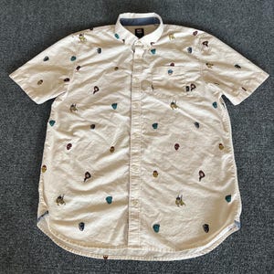 Puede incluir: Camisa abotonada de color crema con mangas cortas, con un estampado de emblemas de superhéroes de colores. La camisa tiene cuello abotonado, bolsillo en el pecho y ribete azul en el cuello interior y los puños.