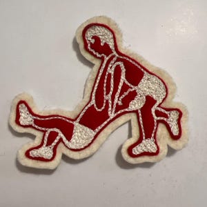 May include: A red felt patch featuring a white embroidered outline of a person in a stretching pose. The figure is depicted in a lunge with one leg extended forward and the other bent, with white fuzzy trim bordering the entire red shape.