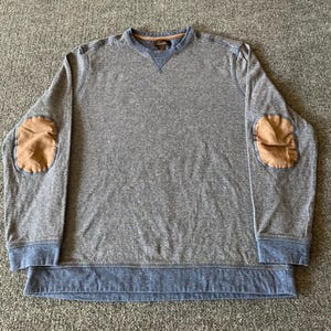 May include: Gray long-sleeve sweatshirt with blue accents and brown elbow patches. The sweatshirt has a crew neck and a ribbed hem. The fabric has a subtle striped pattern. The overall design is casual and comfortable.