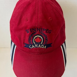 May include: Red baseball cap with the word "WINNIPEG" embroidered in navy blue above a navy blue banner that reads "CANADA". The cap has a maple leaf emblem and white and navy blue stripes on the side.
