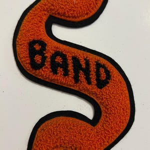 May include: A chenille patch in the shape of a stylized letter S with a bright orange textured surface and a black border. The word BAND is embroidered in black thread across the center of the orange letter S, creating a bold contrast.