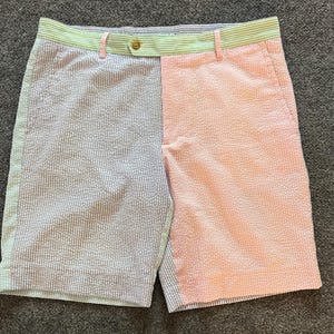May include: Seersucker shorts with a unique color-block design. One side is light blue and white striped, the other is pink and white dotted. The waistband features green and white stripes. A button and a pink belt loop are visible.