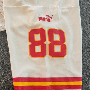 Op de afbeelding: Wit shirt met rode en gele details. Het nummer "88" is rood weergegeven met een gele omlijning. Het Puma-logo is rood. De onderste zoom heeft rode en gele strepen.