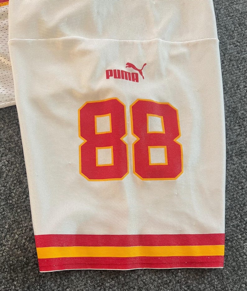 Op de afbeelding: Wit shirt met rode en gele details. Het nummer "88" is rood weergegeven met een gele omlijning. Het Puma-logo is rood. De onderste zoom heeft rode en gele strepen.