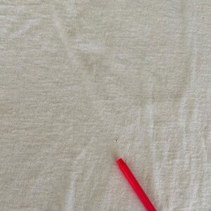 May include: Close-up of a white t-shirt with a gray design at the top. A red pen is in the bottom right corner. The shirt has some small stains.