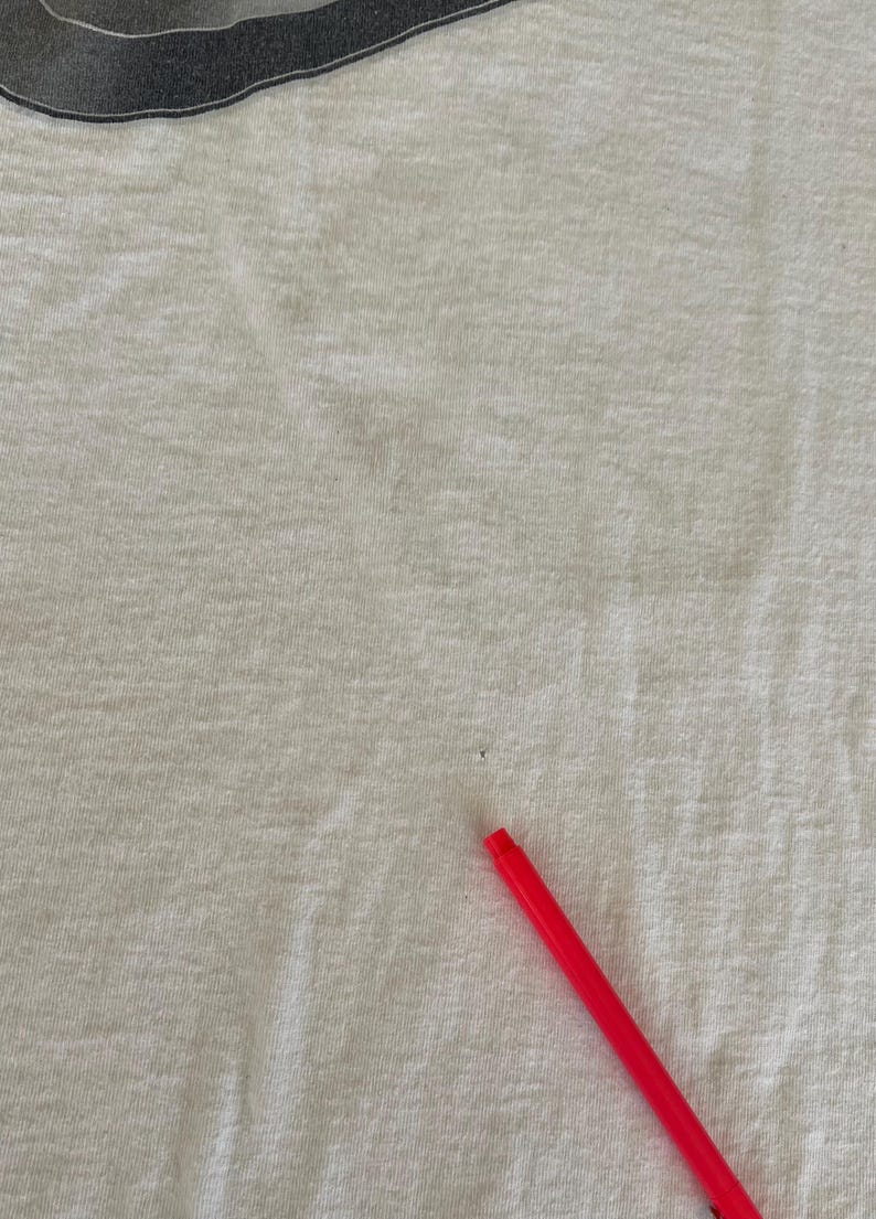 May include: Close-up of a white t-shirt with a gray design at the top. A red pen is in the bottom right corner. The shirt has some small stains.