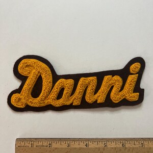 May include: A dark brown felt patch featuring the name Danni embroidered in a textured, golden-yellow cursive script. The patch is positioned above a wooden ruler, showing the design spans approximately 7.25 inches in total length.