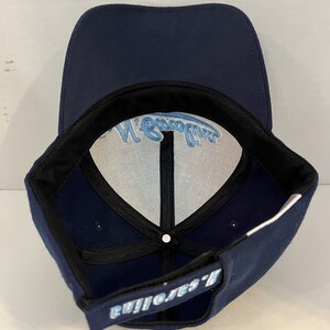 Pu&ograve; includere: Un cappello da baseball blu navy con un logo ricamato bianco e blu chiaro che dice "Eagles' Nest".