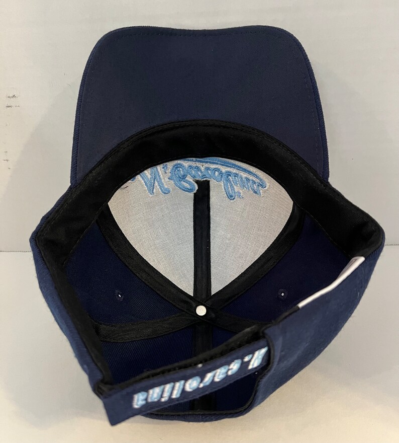 Pu&ograve; includere: Un cappello da baseball blu navy con un logo ricamato bianco e blu chiaro che dice "Eagles' Nest".