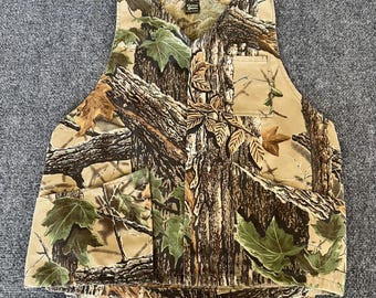Pode incluir: Colete camuflado com um padrão detalhado de árvores e folhas em tons de castanho, verde e bege. O colete inclui vários bolsos e um decote em V. O material parece resistente, adequado para atividades ao ar livre.