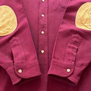 May include: A long-sleeve button-down shirt in a deep burgundy color. The shirt features contrasting tan elbow patches on both sleeves, a central button placket with matching buttons, and buttoned cuffs at the end of each sleeve.