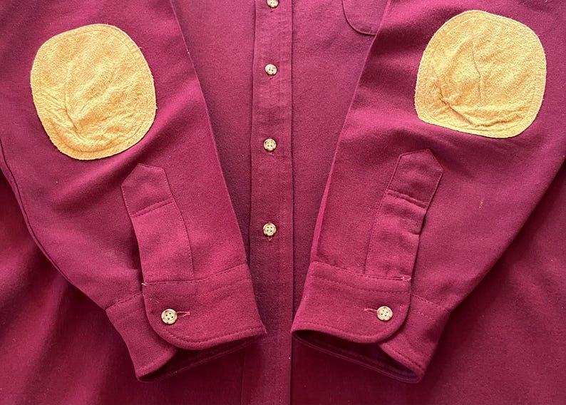 May include: A long-sleeve button-down shirt in a deep burgundy color. The shirt features contrasting tan elbow patches on both sleeves, a central button placket with matching buttons, and buttoned cuffs at the end of each sleeve.