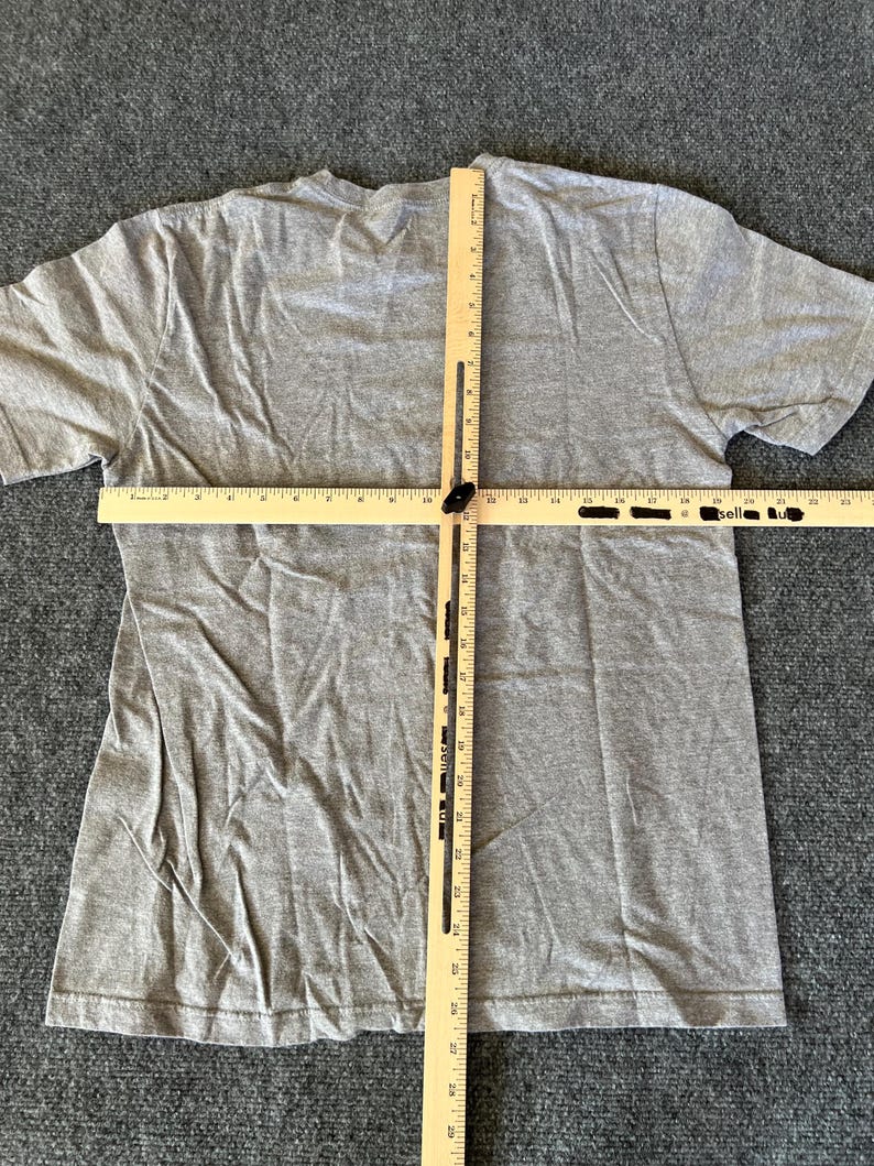 Pu&ograve; includere: Una maglietta grigio melange a maniche corte &egrave; stesa, con un righello di legno posto sul petto e al centro. Le misure della maglietta vengono prese con il righello. La maglietta &egrave; su una superficie grigia.