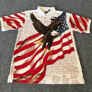 May include: White polo shirt featuring a patriotic design with the American flag and a bald eagle. The shirt's design incorporates the Declaration of Independence text. Red and white stripes and blue stars are visible.