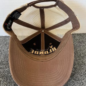 Op de afbeelding: Bruine baseballcap met een mesh achterkant en een gebogen klep. De cap heeft het woord "BRIDE" in goud geborduurd op een zwart paneel. De merknaam "Sportsman" is zichtbaar.