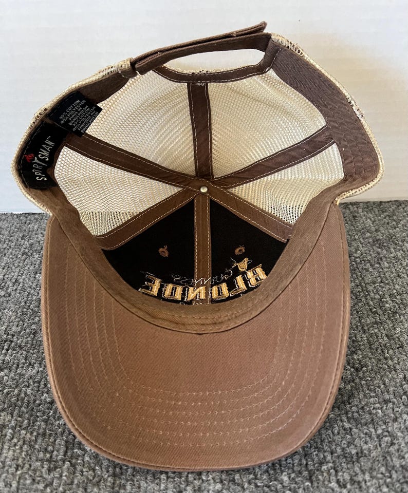 Op de afbeelding: Bruine baseballcap met een mesh achterkant en een gebogen klep. De cap heeft het woord "BRIDE" in goud geborduurd op een zwart paneel. De merknaam "Sportsman" is zichtbaar.