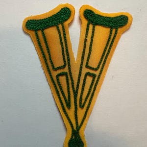 May include: A yellow fabric patch shaped like two crutches joined at the base. The design features dark green embroidered outlines and details, including the armrests and vertical supports, creating a V-shaped silhouette on a smooth yellow background.
