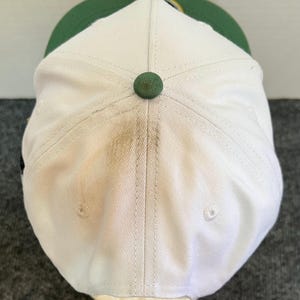 May include: A white baseball cap with a green brim and a green button on top. The cap has a slightly worn appearance, with some discoloration visible on the white fabric. The cap is a classic style, suitable for casual wear.