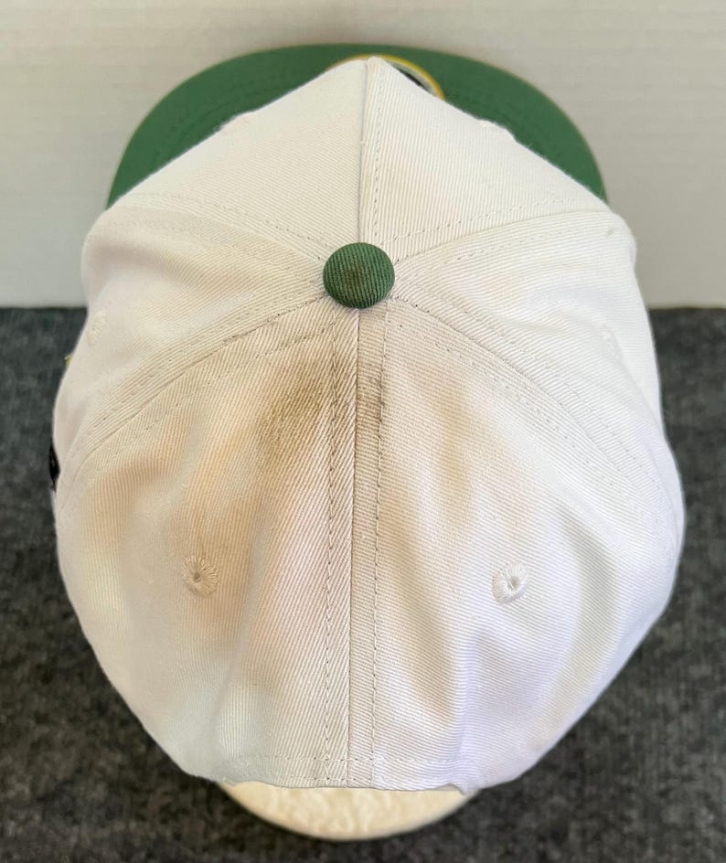 May include: A white baseball cap with a green brim and a green button on top. The cap has a slightly worn appearance, with some discoloration visible on the white fabric. The cap is a classic style, suitable for casual wear.