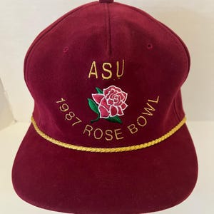 May include: A maroon baseball cap featuring gold embroidered text that reads ASU 1987 ROSE BOWL. The cap includes a central embroidered pink rose with green leaves and a decorative gold braided cord trim running across the base of the front panel.