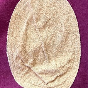 May include: An oval-shaped beige suede elbow patch with a fuzzy, textured surface and visible stitching around the perimeter, securely attached to a deep magenta-colored fabric background with a woven, slightly coarse texture.