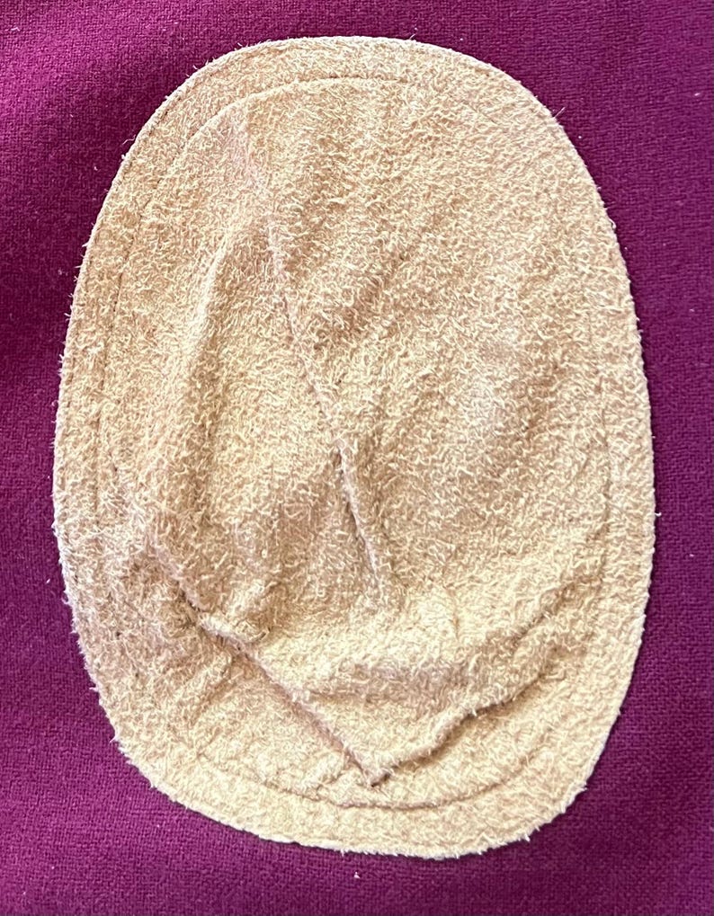 May include: An oval-shaped beige suede elbow patch with a fuzzy, textured surface and visible stitching around the perimeter, securely attached to a deep magenta-colored fabric background with a woven, slightly coarse texture.